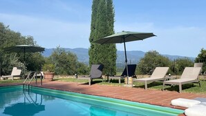 Ein Swimmingpool mit Liegestühlen und Sonnenschirmen, umgeben von Grünflächen und Bergen im Hintergrund.
