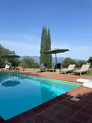 Ein Swimmingpool mit Liegestühlen und Sonnenschirmen, umgeben von Grünflächen und Bergen im Hintergrund.