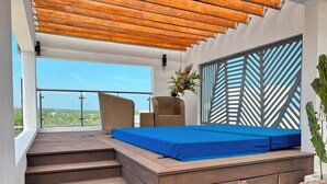 Un balcón con cubierta de madera, un colchón azul y una silla de mimbre.