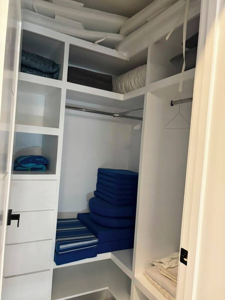 A walk-in closet with multiple shelves and a hanging rod, containing folded clothes and a blue chair.