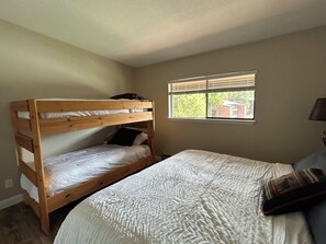 A bedroom with a bunk bed, a single bed, a window with blinds, and a lamp.