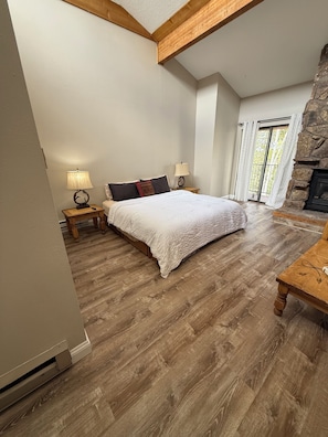 A bedroom with a large bed, wooden flooring, a stone fireplace, and a balcony with curtains.