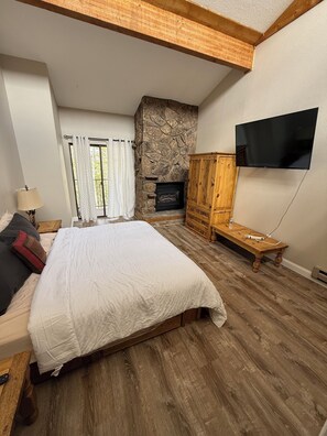 A bedroom with a large bed, a stone fireplace, a wooden wardrobe, a television mounted on the wall, and a window with curtains. - Beautiful 3 Bedroom/2.5 bath Condo Steps from Downtown Winter Park (Winter Park)