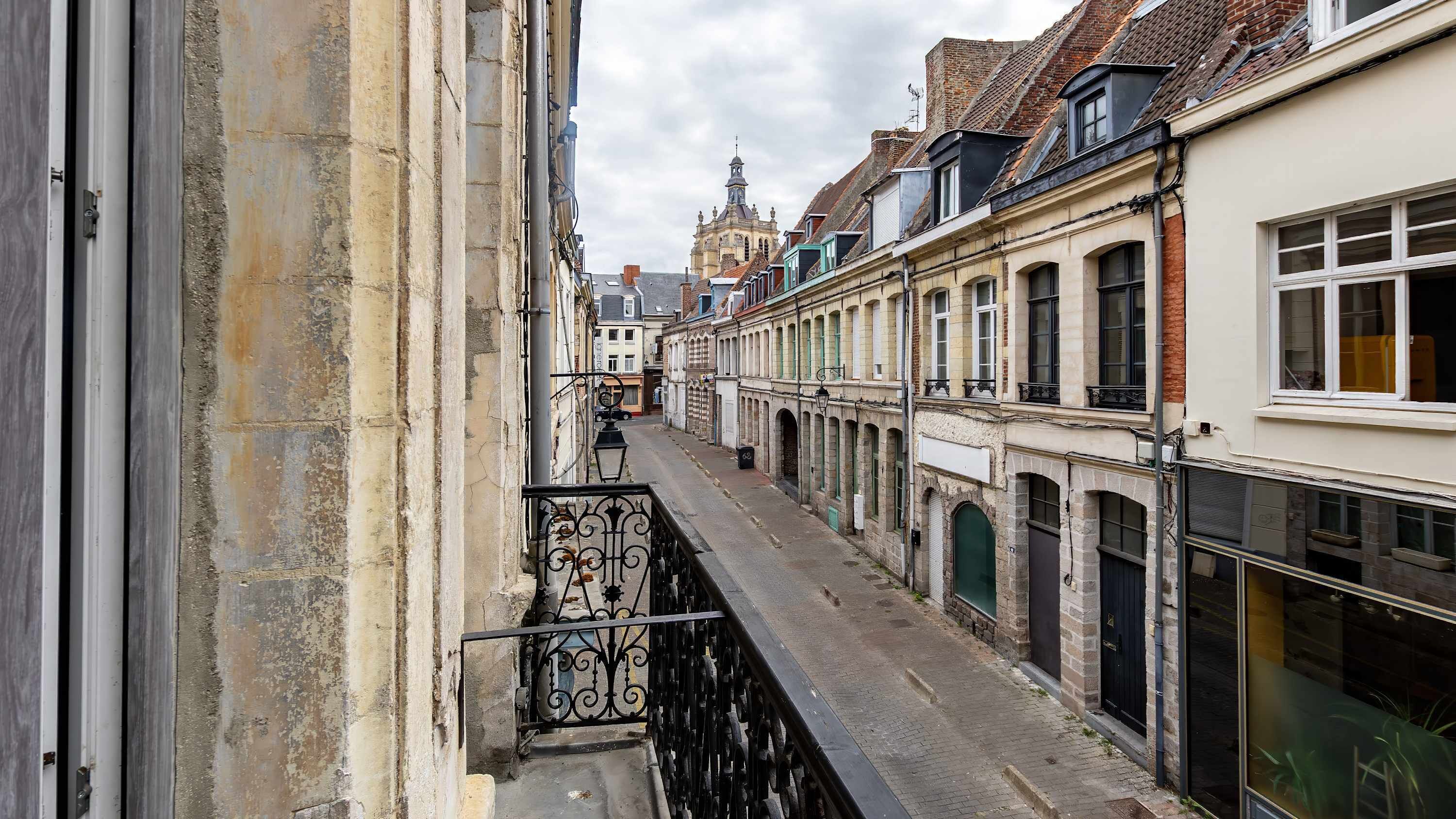 A narrow street with buildings on either side, a visible church spire in the distance, and a metal balcony railing.