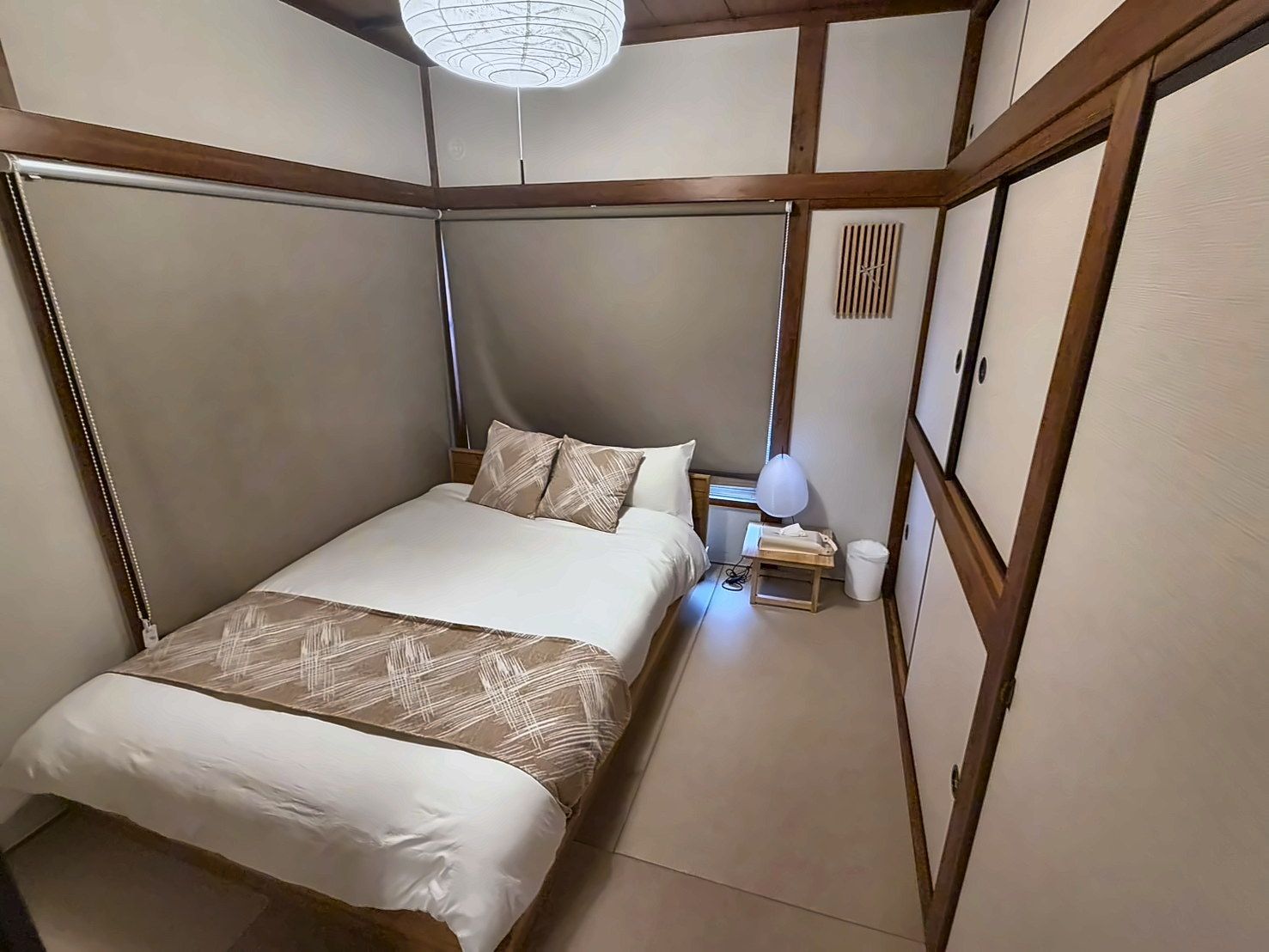 A traditional Japanese-style bedroom with tatami flooring, a low bed with a white and brown bedspread, a bedside table with a lamp, and a shoji screen door.