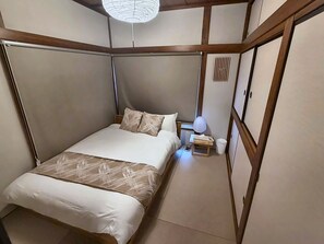 A traditional Japanese-style bedroom with tatami flooring, a low bed with a white and brown bedspread, a bedside table with a lamp, and a shoji screen door. - MSTAY Oshiage One (Sumida-ku)