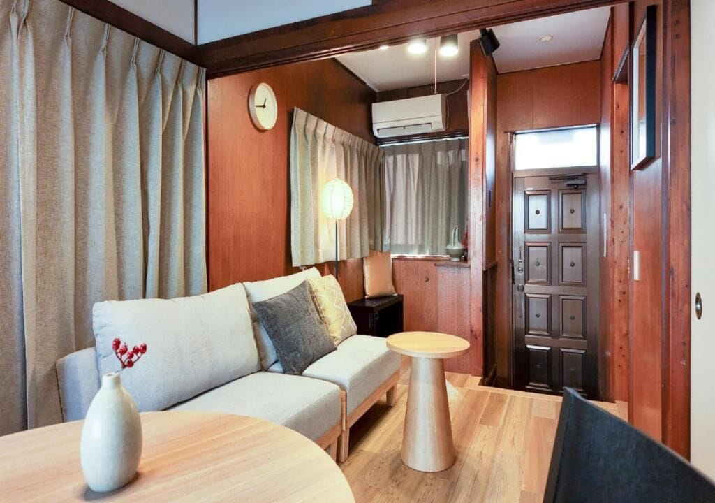 A room with a sofa, a round wooden table, a vase with flowers, a wall clock, and an air conditioning unit.