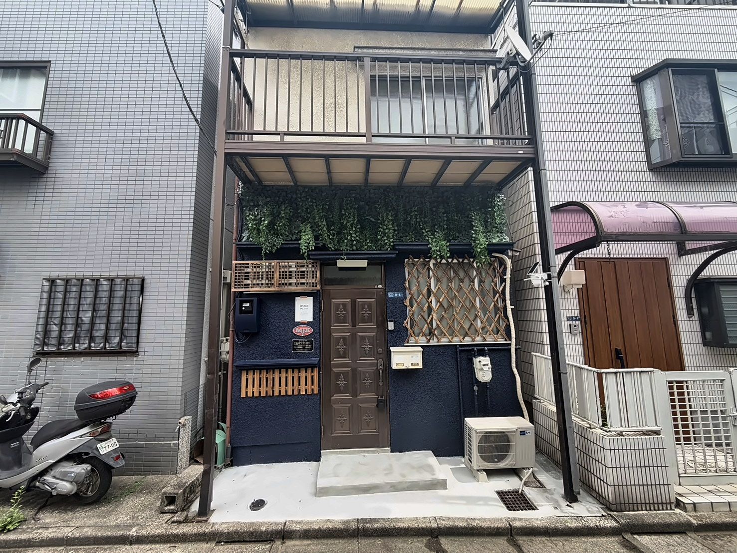 A two-story building with a gated entrance, a scooter parked outside, and an air conditioning unit on the ground floor.