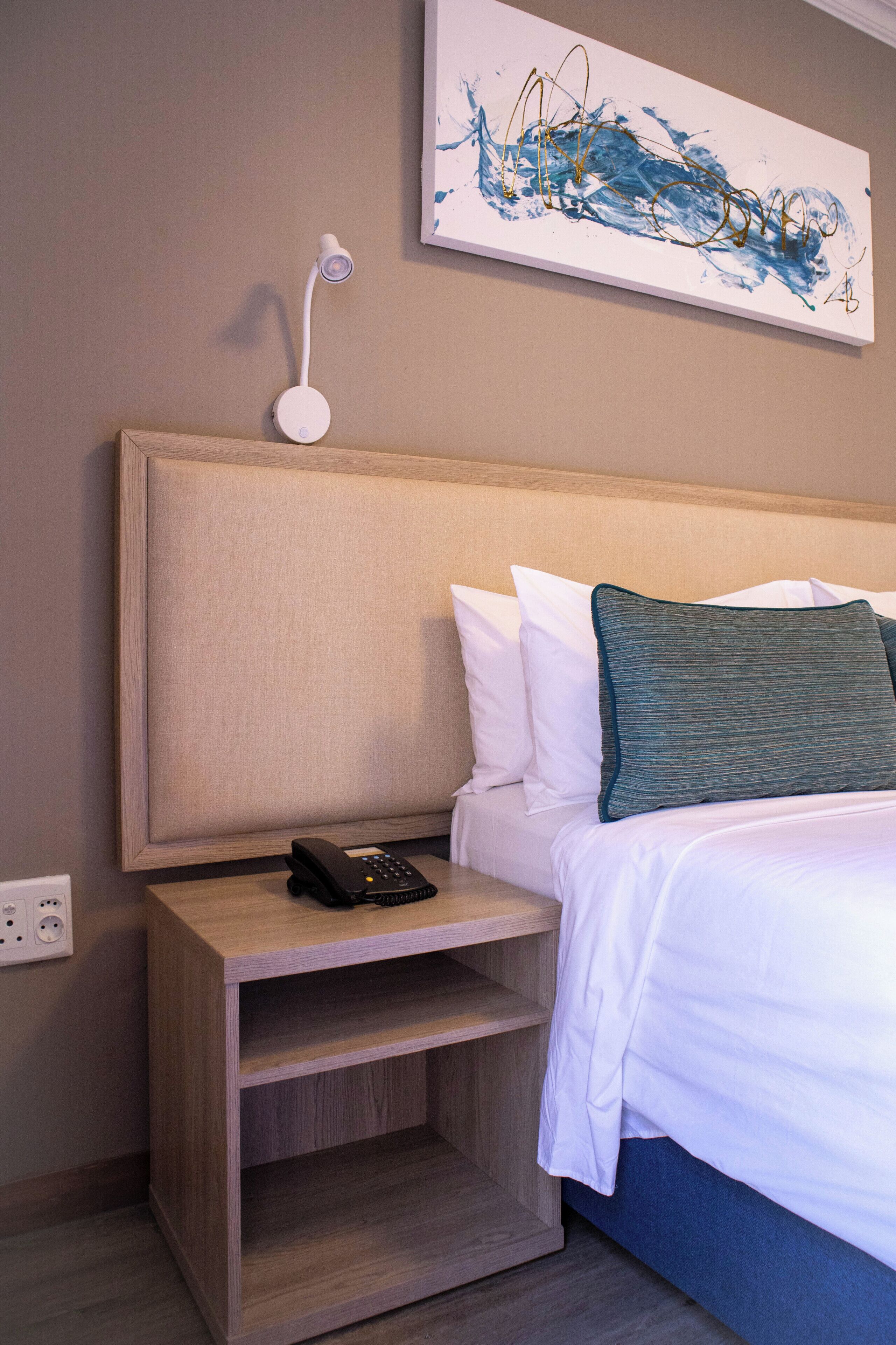 Habitación de hotel con cama, mesita de noche con teléfono, lámpara de pared y un cuadro.