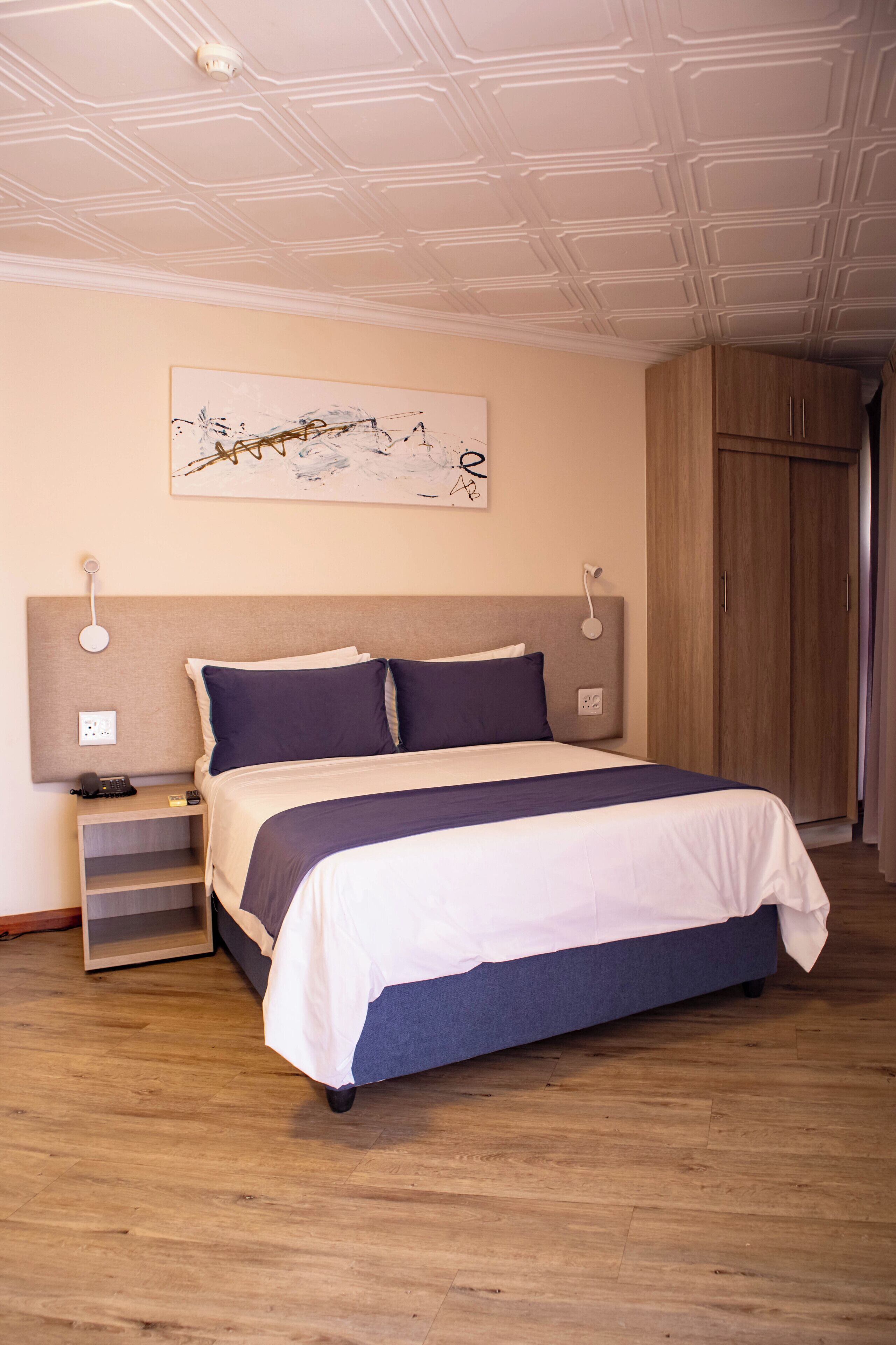 Un dormitorio con una cama, mesitas de noche, una lámpara de pared, techo con un diseño geométrico y un armario de madera.