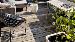 Una terrazza esterna con pavimento in legno, un'area salotto con cuscini, un tavolo con sedie e piante in vaso.