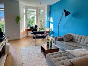 Ein Wohnzimmer mit einer Couch, einem Couchtisch, einem Schreibtisch mit Computer und einer hängenden Pflanze.