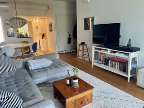 A modern living room with a grey sofa, a wooden coffee table with a bottle and plant, a TV on a white stand, and a bookshelf with books and a board game. - Designer Light Filled 2BR House in Kensington w/Rooftop patio (Toronto)
