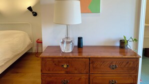 Una camera da letto con un letto, una cassettiera in legno, una lampada, una piccola pianta in vaso, una lampada a parete e un quadro astratto dai colori vivaci.