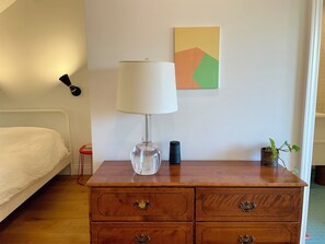 Ein Schlafzimmer mit einem Bett, einer hölzernen Kommode mit Schubladen, einer Lampe, einem kleinen Topfpflanzen und einem Wandleuchter, sowie einem farbenfrohen abstrakten Gemälde.