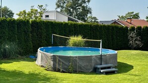 Piscina subterrânea circular com uma rede, rodeada por um relvado bem cuidado, com um sequeiro e edifícios ao fundo.
