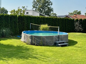 Una piscina elevada circular con una red, rodeada por un césped cuidado, con setos y edificios al fondo.