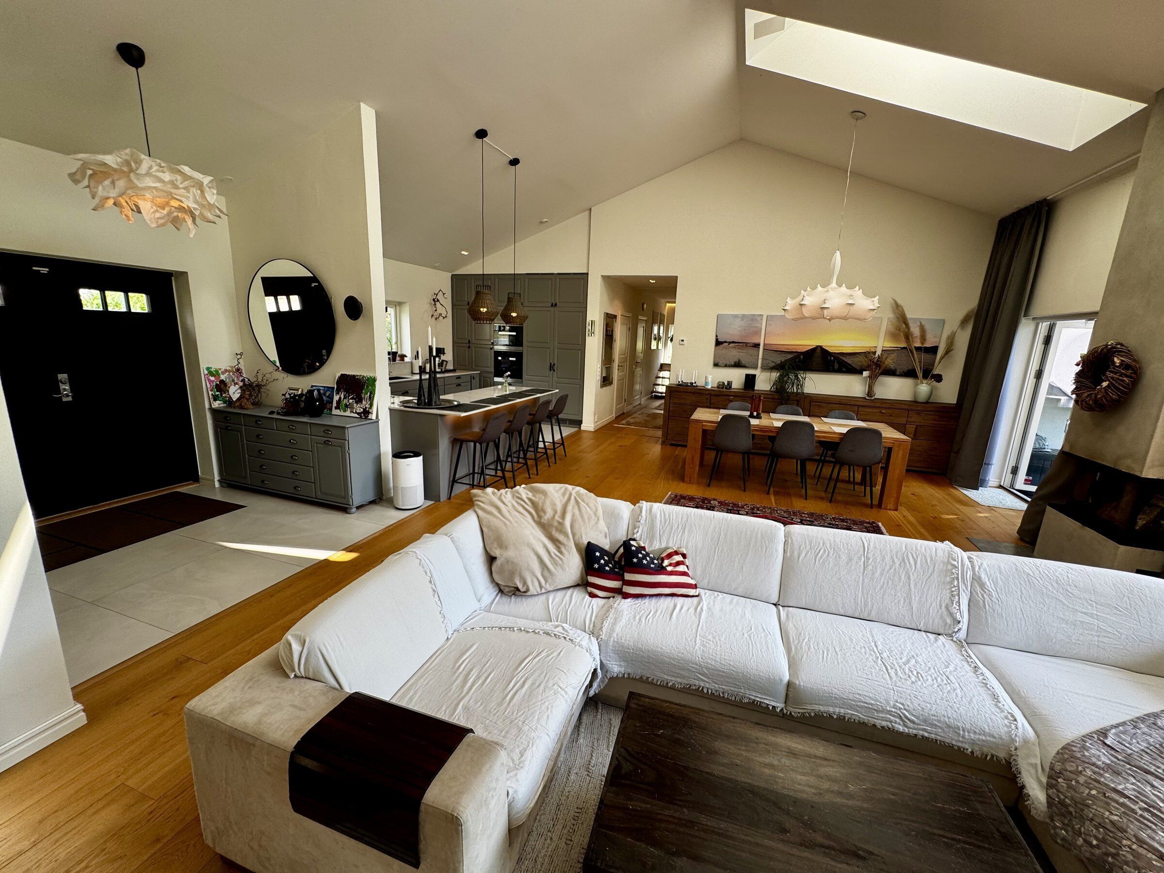 Un salón amplio con un sofá seccional blanco grande, una mesa de comedor con sillas, una cocina con isla central y un techo con tragaluz.