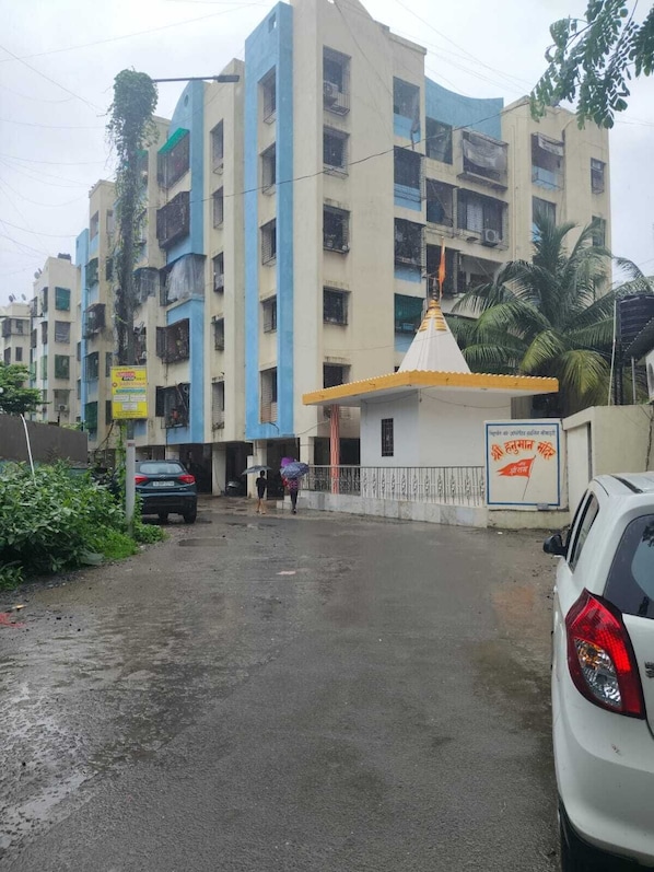 A multi-story building with a gated entrance, a small shrine-like structure, and parked cars.