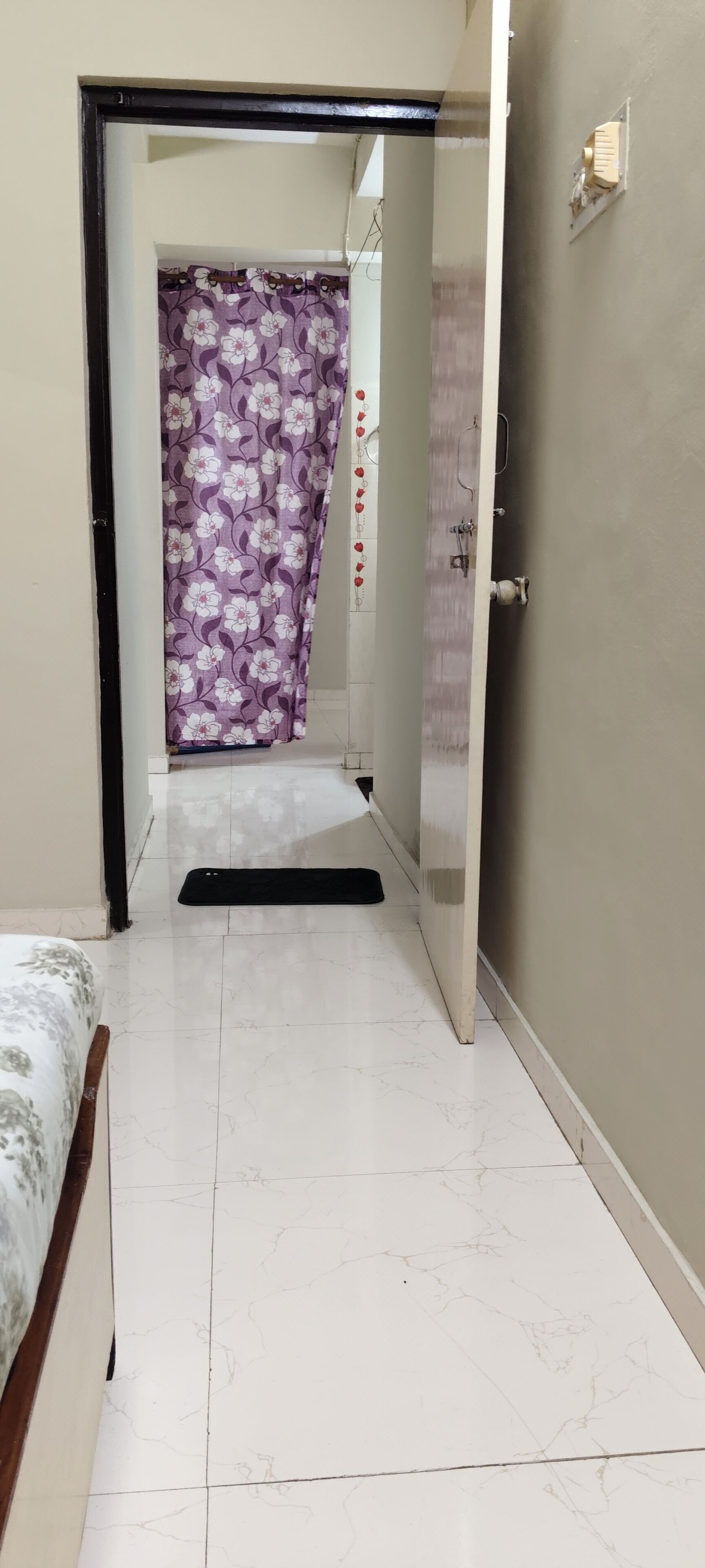 Um corredor com uma cama, uma porta e um banheiro com cortina florida.