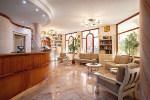 A hotel lobby with a reception desk, seating area, bookshelves, and large windows.