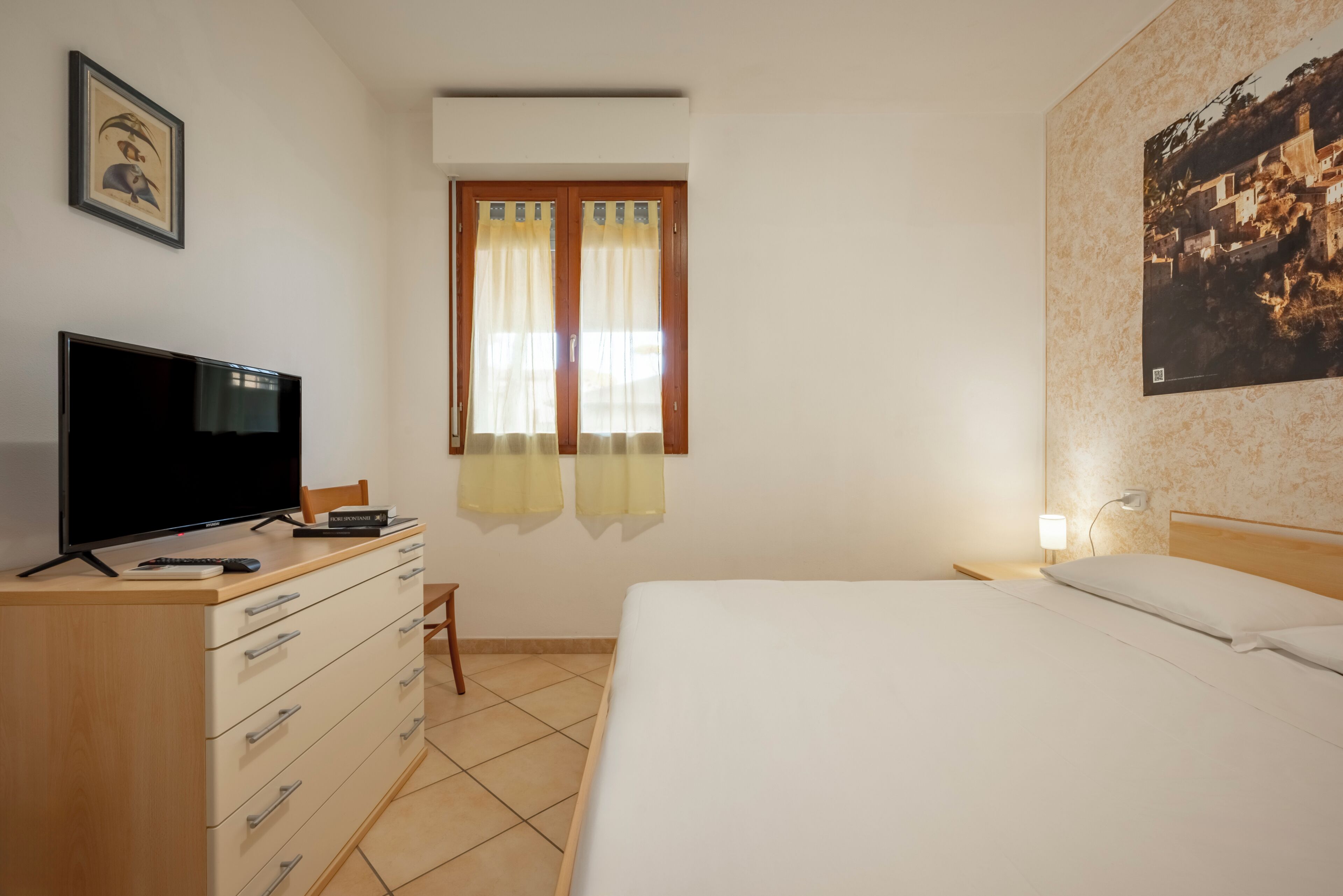 Una camera d'albergo con un letto, una televisione, una scrivania con una sedia, una finestra con tende e un quadro appeso al muro.