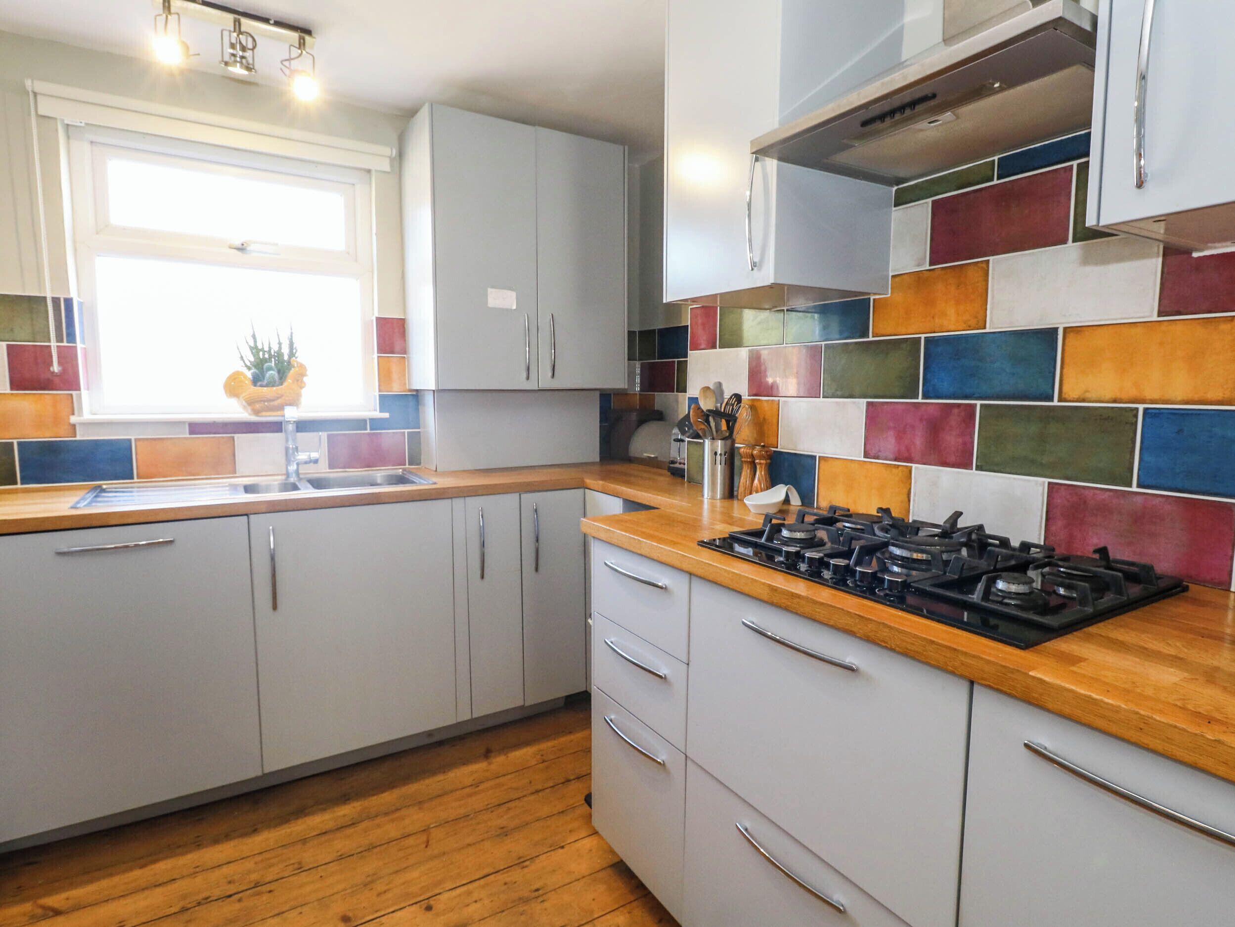 A kitchen with grey cabinets, a wooden countertop, a colorful backsplash, and a window with natural light.