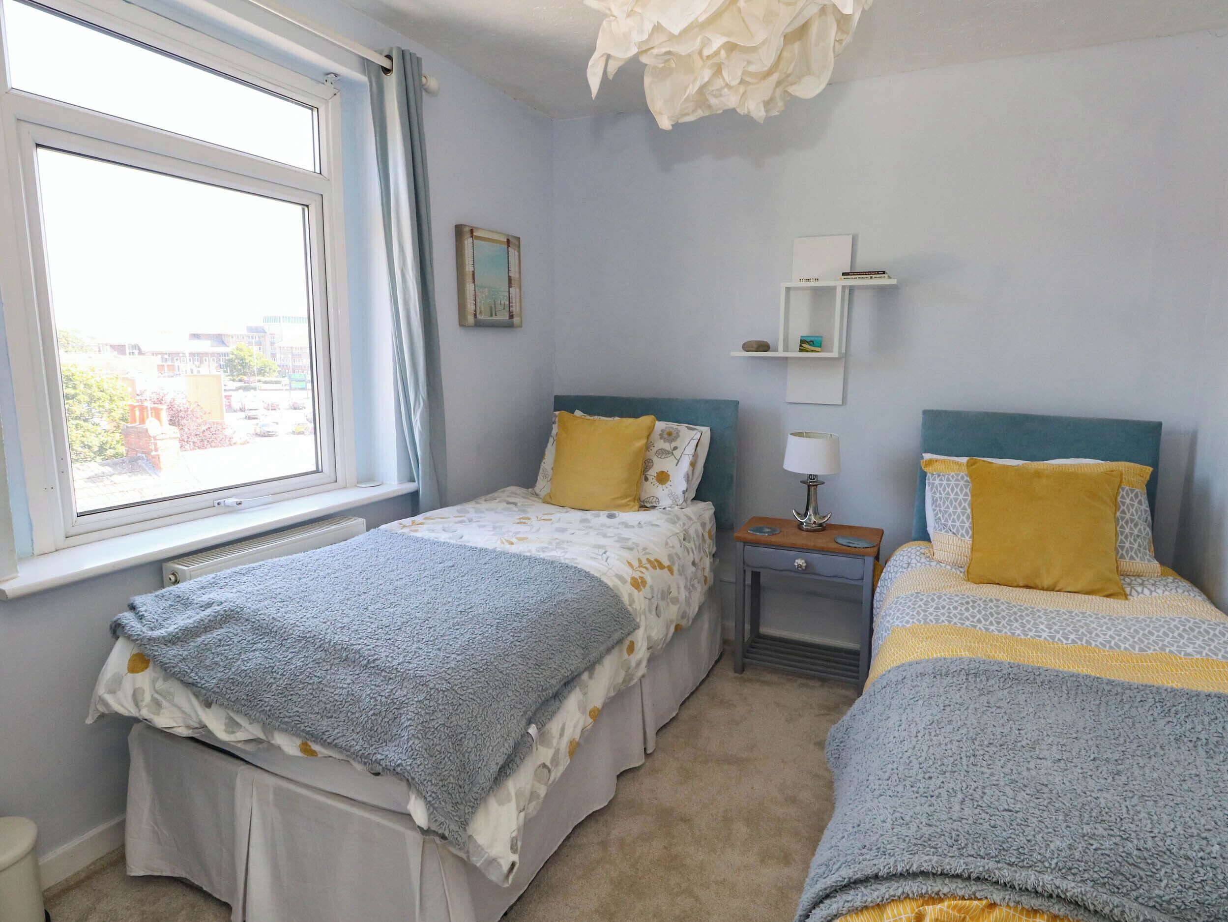 A bedroom with two beds, a window, a radiator, a bedside table with a lamp, and a shelf with a picture.