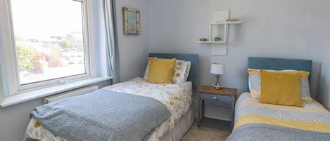 Un dormitorio con dos camas, una ventana, un radiador, una mesita de noche con una lámpara y una estantería con un cuadro.