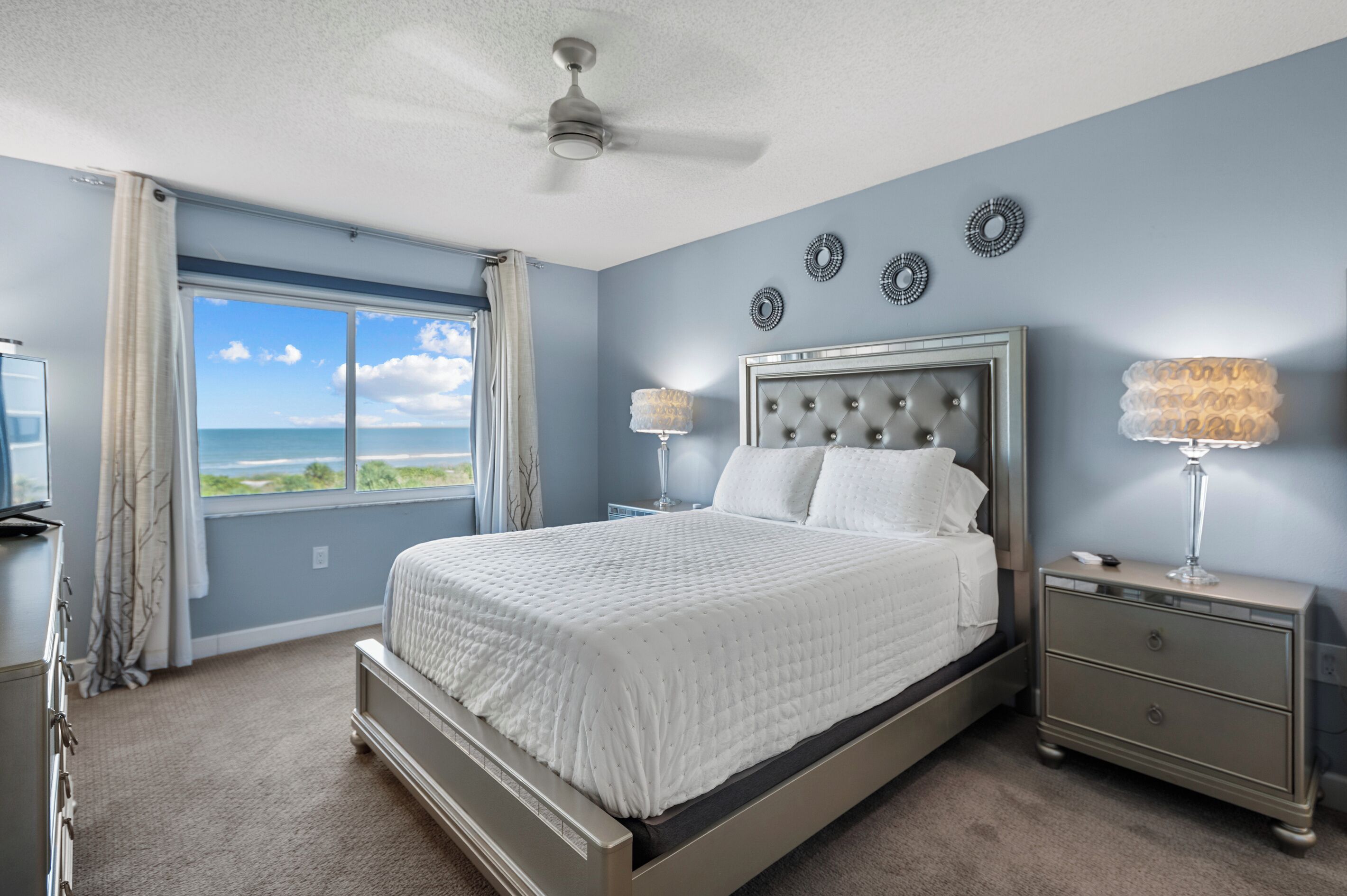 A bedroom with a bed, nightstands, a ceiling fan, a window with a view, and wall decor.