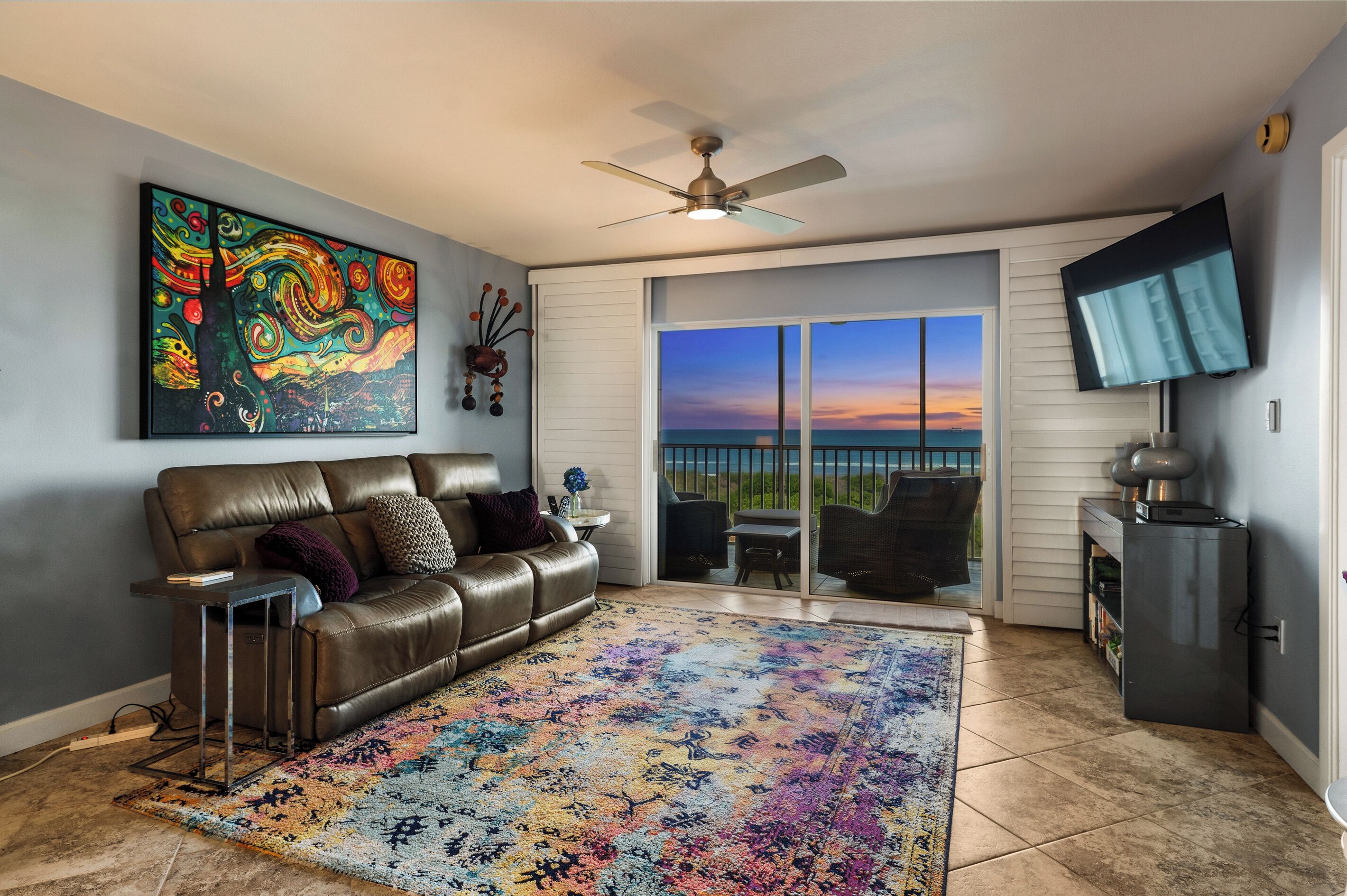 A living room with a leather couch, a colorful rug, a ceiling fan, and a view of the ocean through a sliding glass door.
