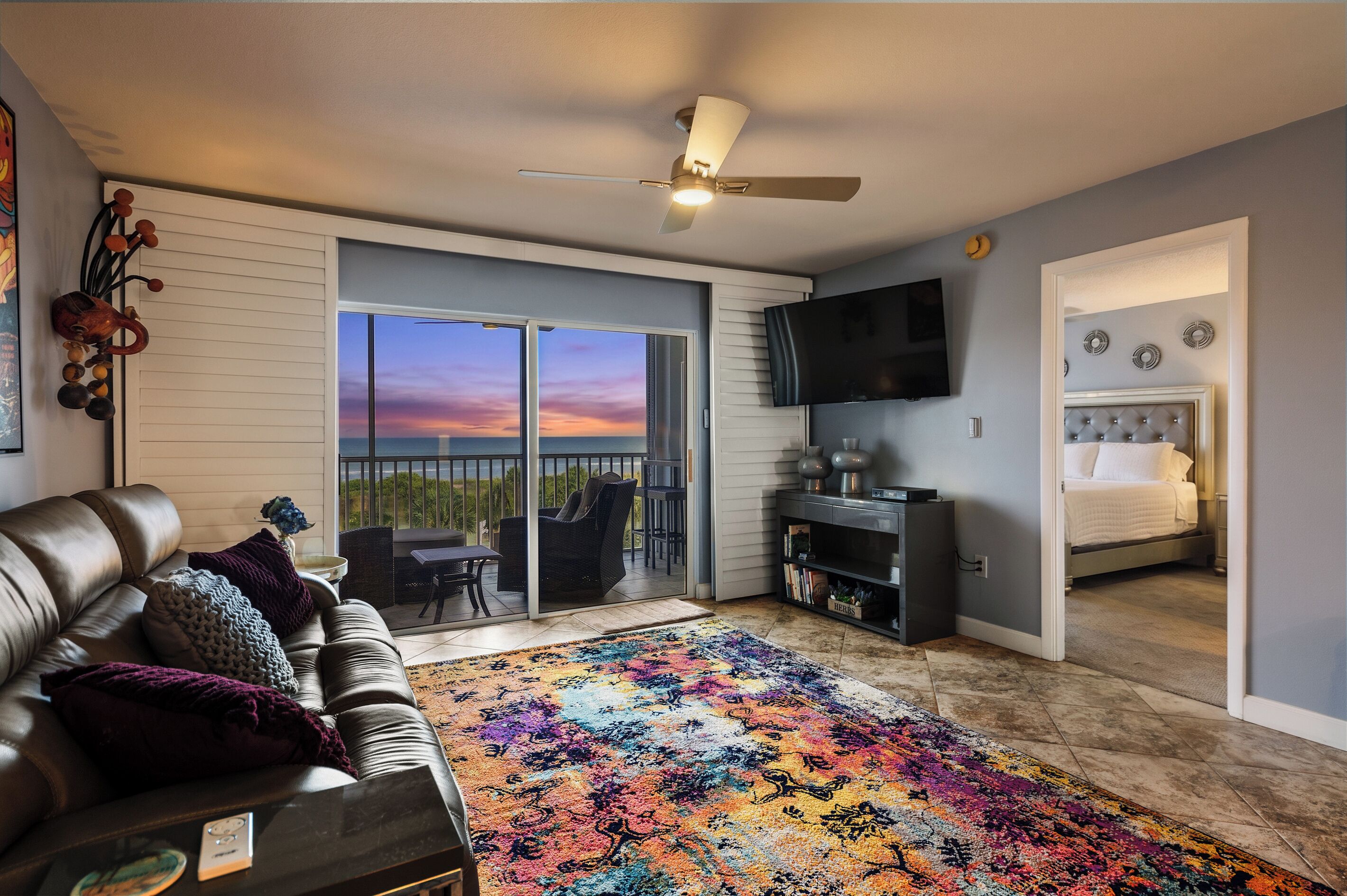 Ein modernes Wohnzimmer mit einer Ledersofa, einem farbenfrohen Teppich, einem Flachbildfernseher und einem Balkon mit Sonnenuntergangsblick.