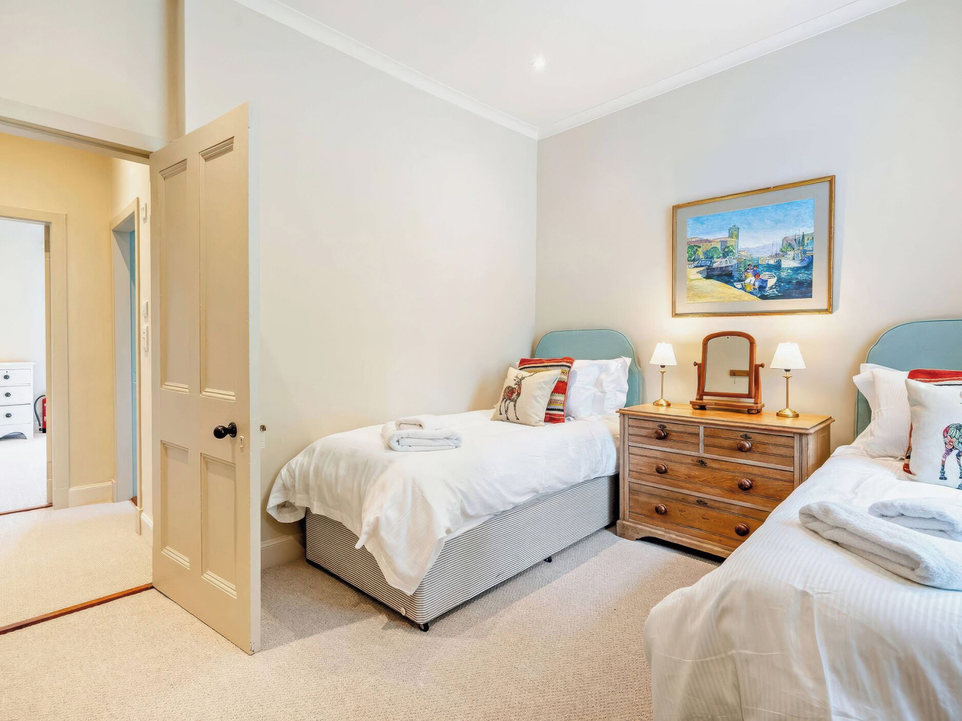 A hotel room with two beds, a dresser, a painting on the wall, and a door leading to another room.