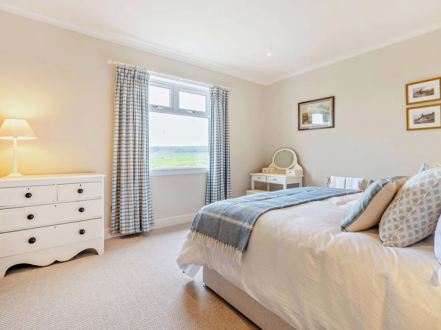 A bedroom with a bed, a dresser, a nightstand, a window with curtains, and a wall with framed pictures.