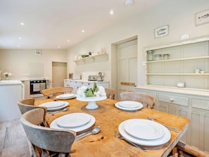 Comedor con mesa de madera dispuesta para seis personas, rodeada de sillas, en una cocina con armarios blancos y un microondas.