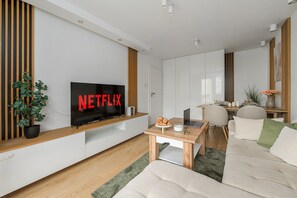 Un saló modern amb un televisor de pantalla plana que mostra 'NETFLIX' en lletres vermelles, una taula de centre de fusta, un sofà, una zona de menjador amb una taula i cadires, i una planta en test.