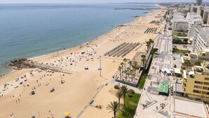 A coastal cityscape with a wide sandy beach, a promenade lined with palm trees, and modern buildings. - Gira Sol by Hdproperties (Quarteira)