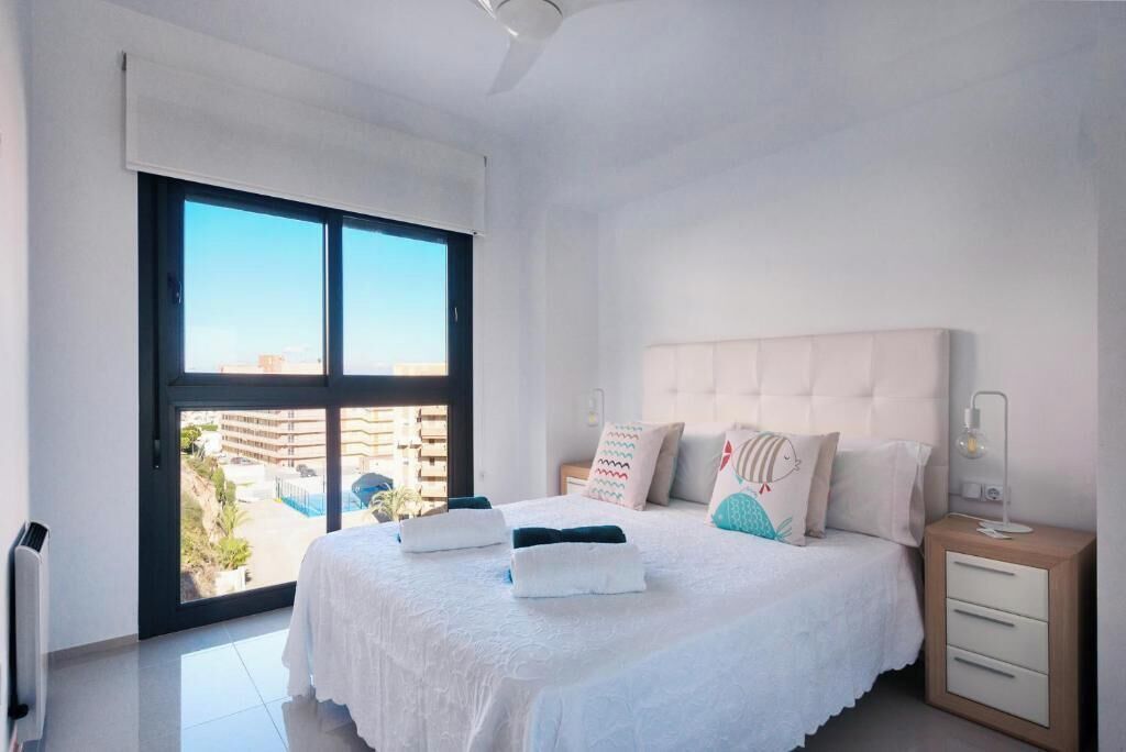Un dormitorio con un ventanal grande, una cama con almohadas, una mesita de noche y un velador.