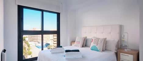 Un dormitorio con un ventanal grande, una cama con almohadas, una mesita de noche y un velador.