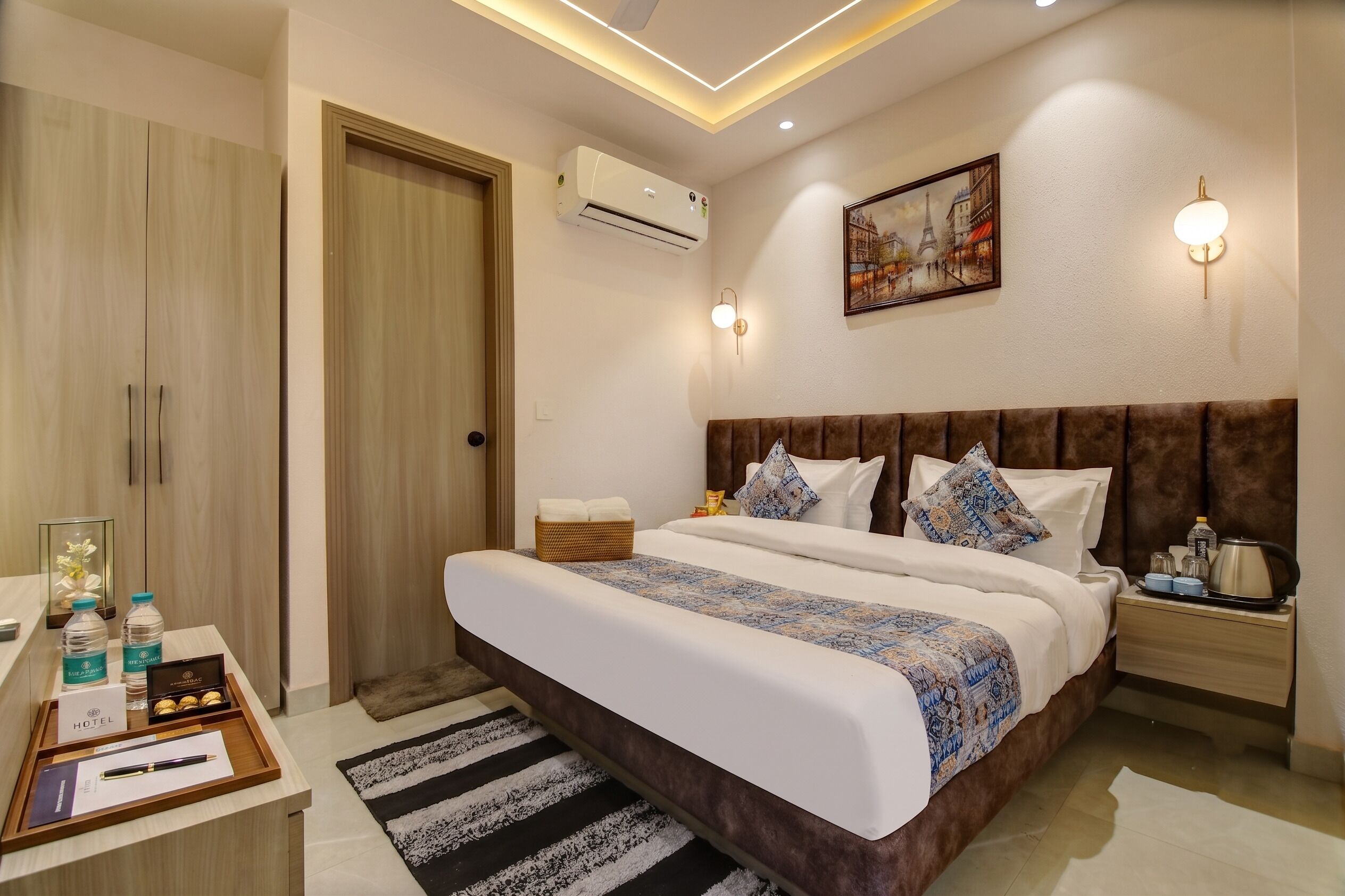 Una habitación de hotel con una cama grande, un armario, una mesita de noche con artículos de aseo, un cuadro en la pared y un equipo de aire acondicionado.