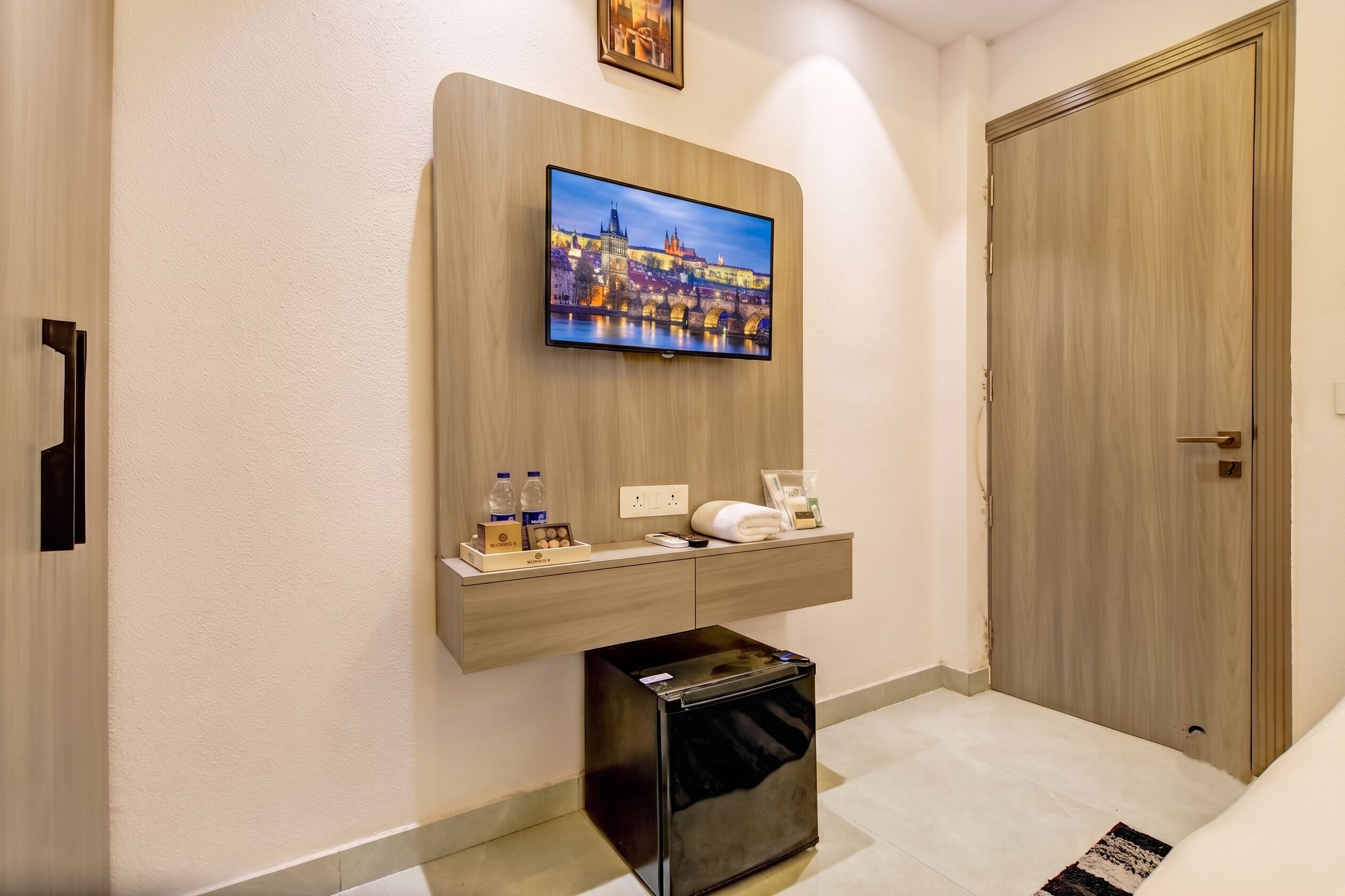 Habitación de hotel con televisión de pantalla plana montada en un panel de madera, un bote de basura pequeño, una toalla y un minibar con agua embotellada y aperitivos.