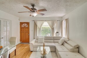 Una sala de estar con una mesa de centro de vidrio, un sofá seccional color beige, un ventilador de techo y una puerta de madera.