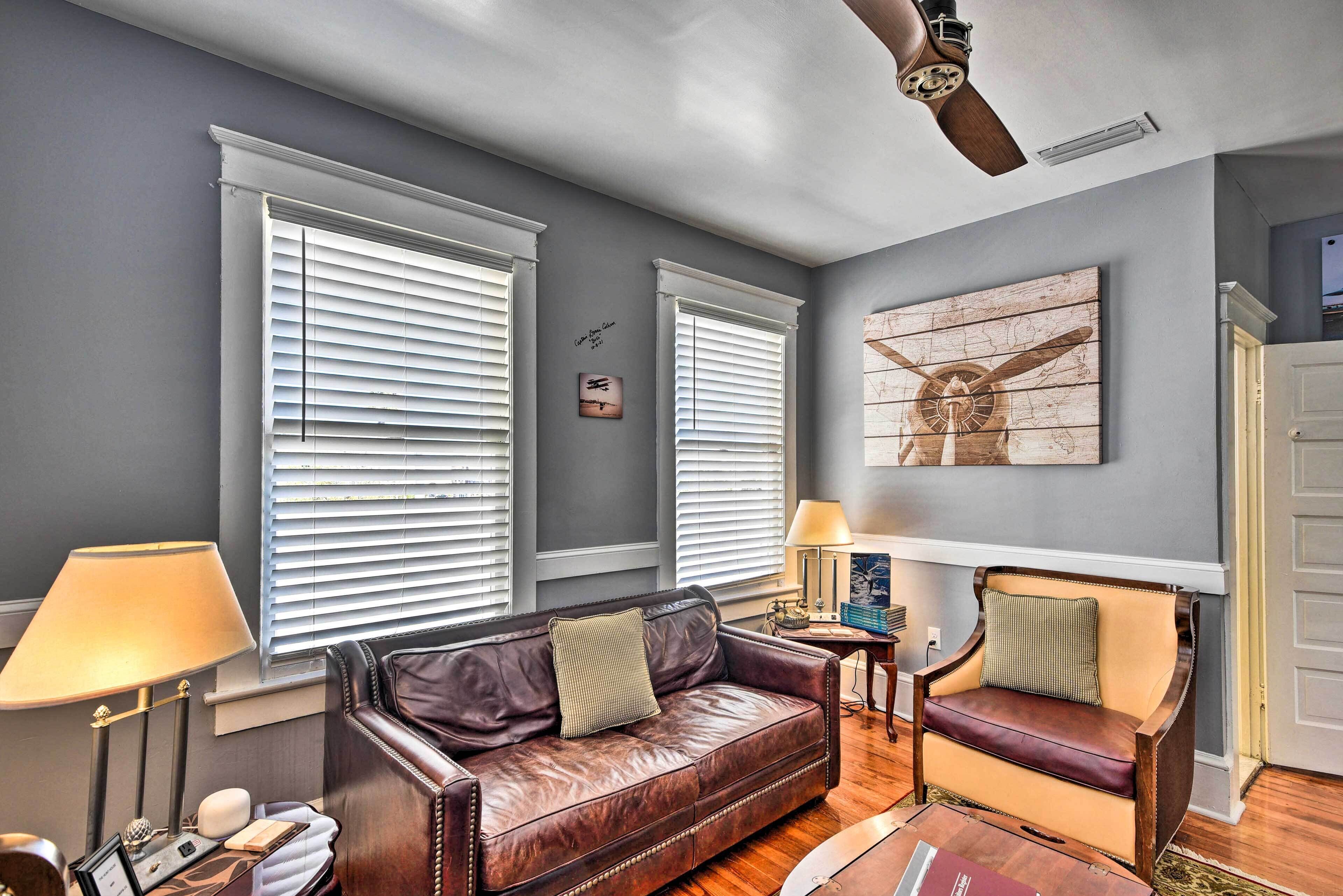 A living room with a leather sofa, two armchairs, a ceiling fan, and a large window with blinds.