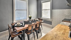 Comedor con mesa y sillas de madera, dos ventanas con persianas, y una zona de cocina con salpicadero de azulejos.