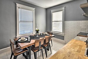 Ein Essbereich mit Holztisch und Stühlen, zwei Fenster mit Jalousien sowie eine Küchenzeile mit gefliestem Spritzschutz.