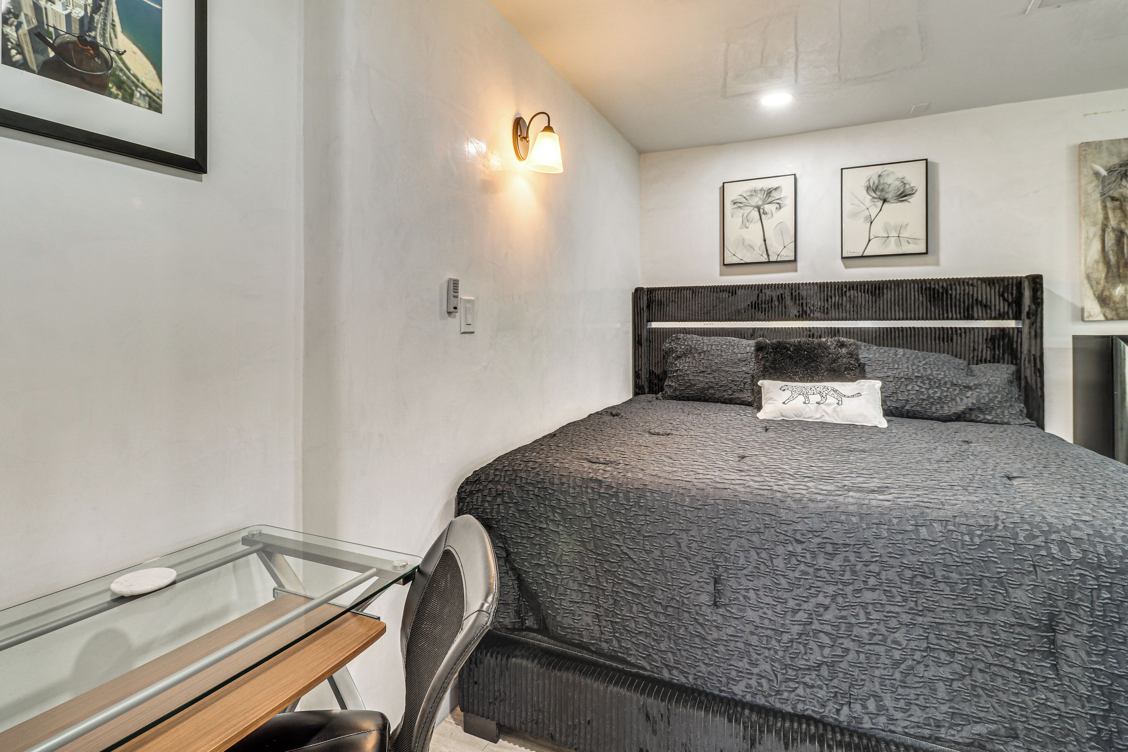 A bedroom with a bed, a bedside table, a chair, a wall-mounted light, and framed artwork.