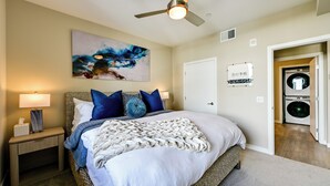 A bedroom with a bed, a nightstand, a ceiling fan, a wall-mounted artwork, a door, and a washing machine in the adjacent room. - Steps to the Sand! Luxury Oceanside Pier Condo (Oceanside)