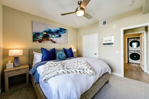 A bedroom with a bed, a nightstand, a ceiling fan, a wall-mounted artwork, a door, and a washing machine in the adjacent room. - Steps to the Sand! Luxury Oceanside Pier Condo (Oceanside)