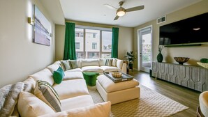 A modern living room with a sectional sofa, a ceiling fan, a flat-screen TV, and a view of the outdoors through large windows. - Steps to the Sand! Luxury Oceanside Pier Condo (Oceanside)