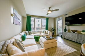 A modern living room with a sectional sofa, a ceiling fan, a flat-screen TV, and a view of the outdoors through large windows. - Steps to the Sand! Luxury Oceanside Pier Condo (Oceanside)